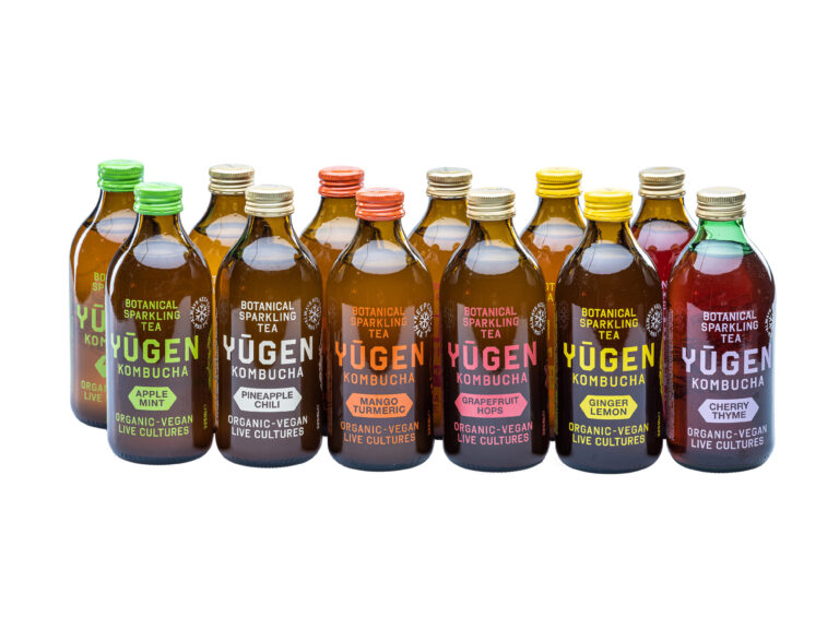 Row of Yugen Kombucha bottles with various flavors including Apple Mint, Pineapple Chili, Mango Turmeric, Grapefruit Hops, Ginger Lemon, and Cherry Thyme, labeled as organic and vegan with live cultures.